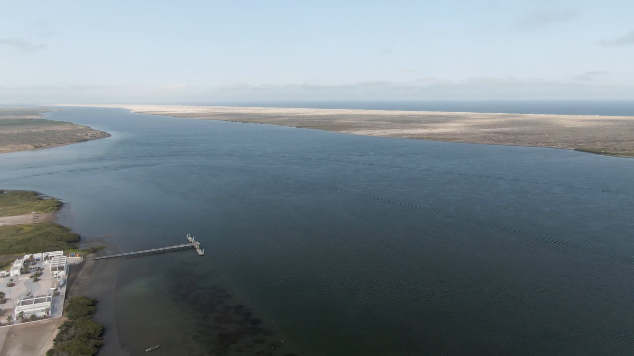 Aerial coastal water scene with dock, shoreline buildings, and distant low land under light clouds.