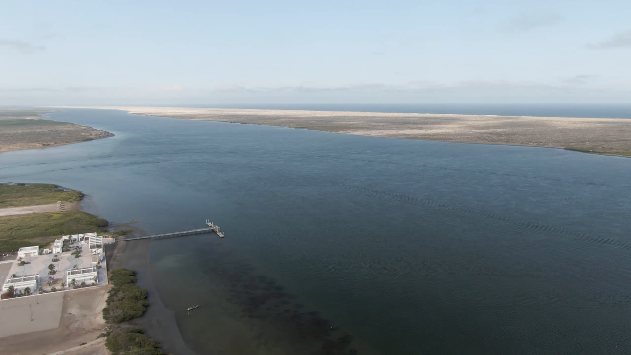 Aerial view in Aldofo Lopez Mateos, Baja California Sur, with a pale shoreline building at lower left, a dock extending into blue-green water, distant sandbars, and no visible people.