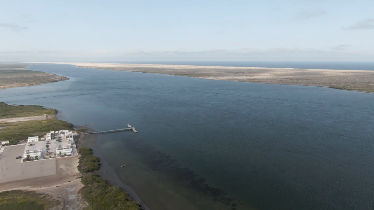 Drone-style coastal water view with dock, building, and distant sandbars.