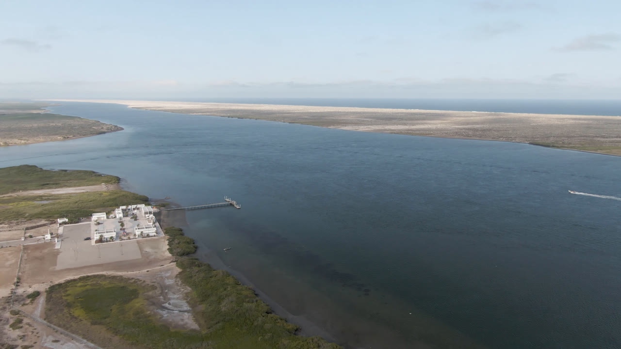 Opening aerial frame over Aldofo Lopez Mateos, Baja California Sur, with a waterfront facility at lower left, broad blue-green water through the center, pale sandbars in the distance, and no visible people.