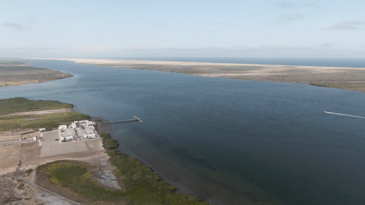 One second into the clip, the Aldofo Lopez Mateos aerial view shows the shoreline facility lower left, the channel widening across the frame, a small white wake on the water, and no visible people.