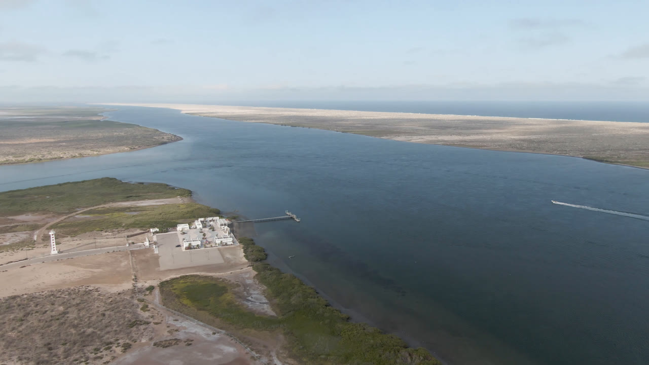 Wide aerial view of a blue water channel, shoreline scrub, dock, and waterfront buildings.