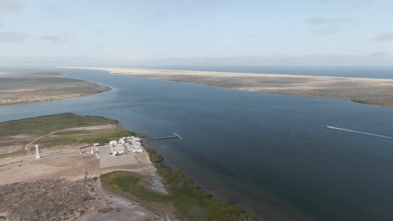 Aerial view in Aldofo Lopez Mateos showing the water channel with a bright boat wake farther across the frame, low scrubland, dock infrastructure, waterfront structures, and no visible people.