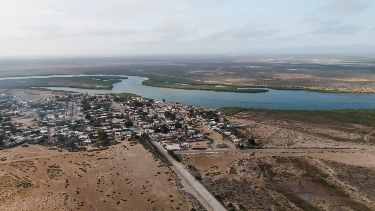 Aerial frame over Adolfo Lopez Mateos, Mexico, showing low buildings clustered near sandy roads, a broad blue river channel, flat land beyond, and no visible people.