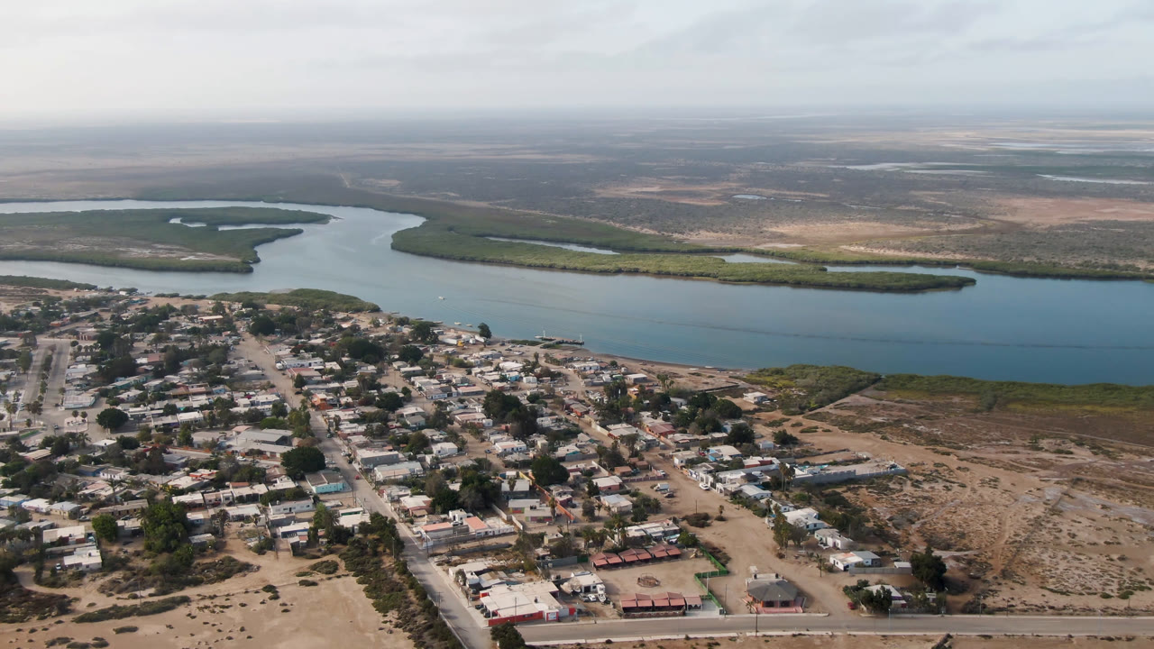 Opening aerial frame over Adolfo Lopez Mateos, Mexico, with low-rise buildings and unpaved roads below a wide river, mangrove islands, dry flats, hazy sky, and no visible people.