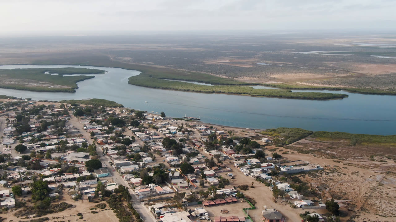 Aerial frame over Adolfo Lopez Mateos, Mexico, with the village grid lower in frame, the blue river running across the middle, mangroves on the far bank, and no visible people.