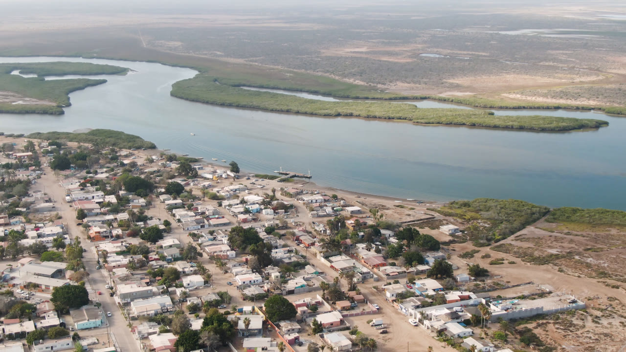 Aerial frame over Adolfo Lopez Mateos, Mexico, with low homes and unpaved roads below, a broad river across the center, mangroves beyond the water, and no visible people.