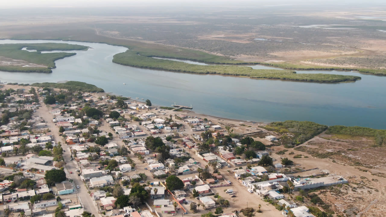 Aerial view of Adolfo Lopez Mateos, Mexico, showing denser village blocks in the lower left, open sandy lots to the right, the river in the midground, and no visible people.