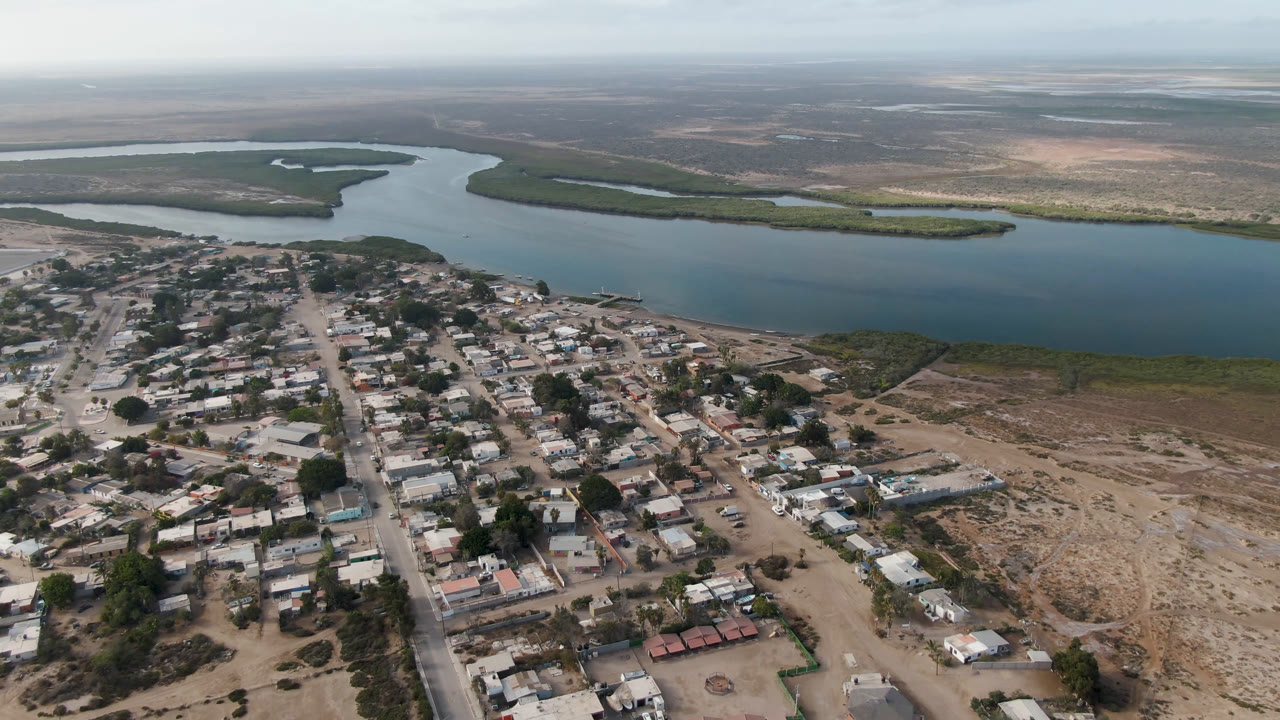 Aerial frame over Aldofo Lopez Mateos showing a central road through low rooftops, scattered trees, a wide blue water channel, green shoreline strips, distant dry terrain, and no visible people.
