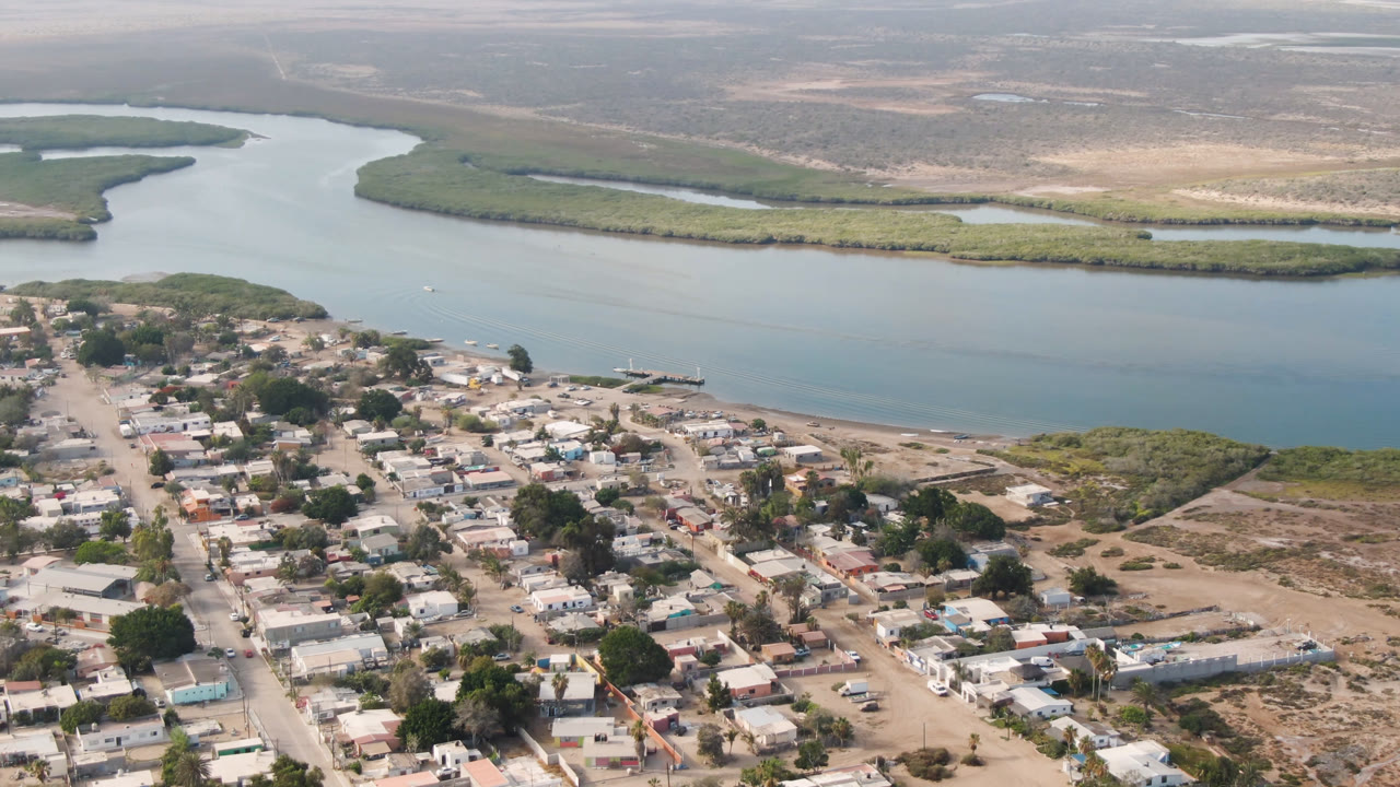 Aerial frame over Aldofo Lopez Mateos, Baja California Sur, with low buildings and dusty roads below, a wide river channel across the middle, marsh islands beyond, and no visible people.