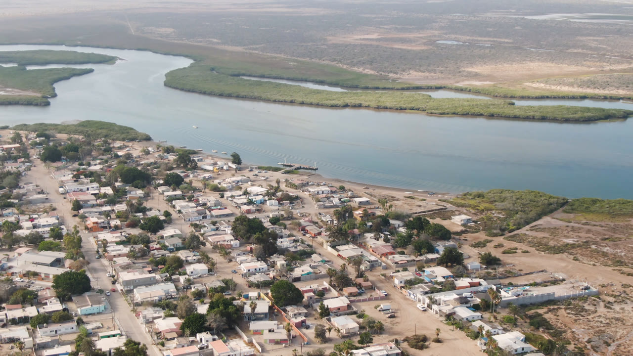 Drone view of Aldofo Lopez Mateos with clustered homes in the foreground, a pale beach strip along the river, green marshland across the water, and no visible people.