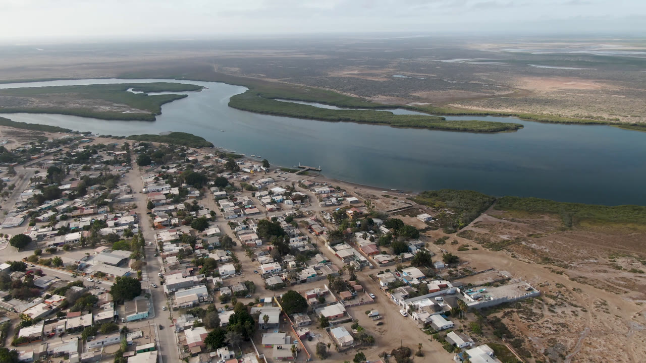 Aerial frame over Aldofo Lopez Mateos showing the town grid slightly farther across the foreground, a curved blue water channel in the midground, vegetated shoreline strips, and no visible people.