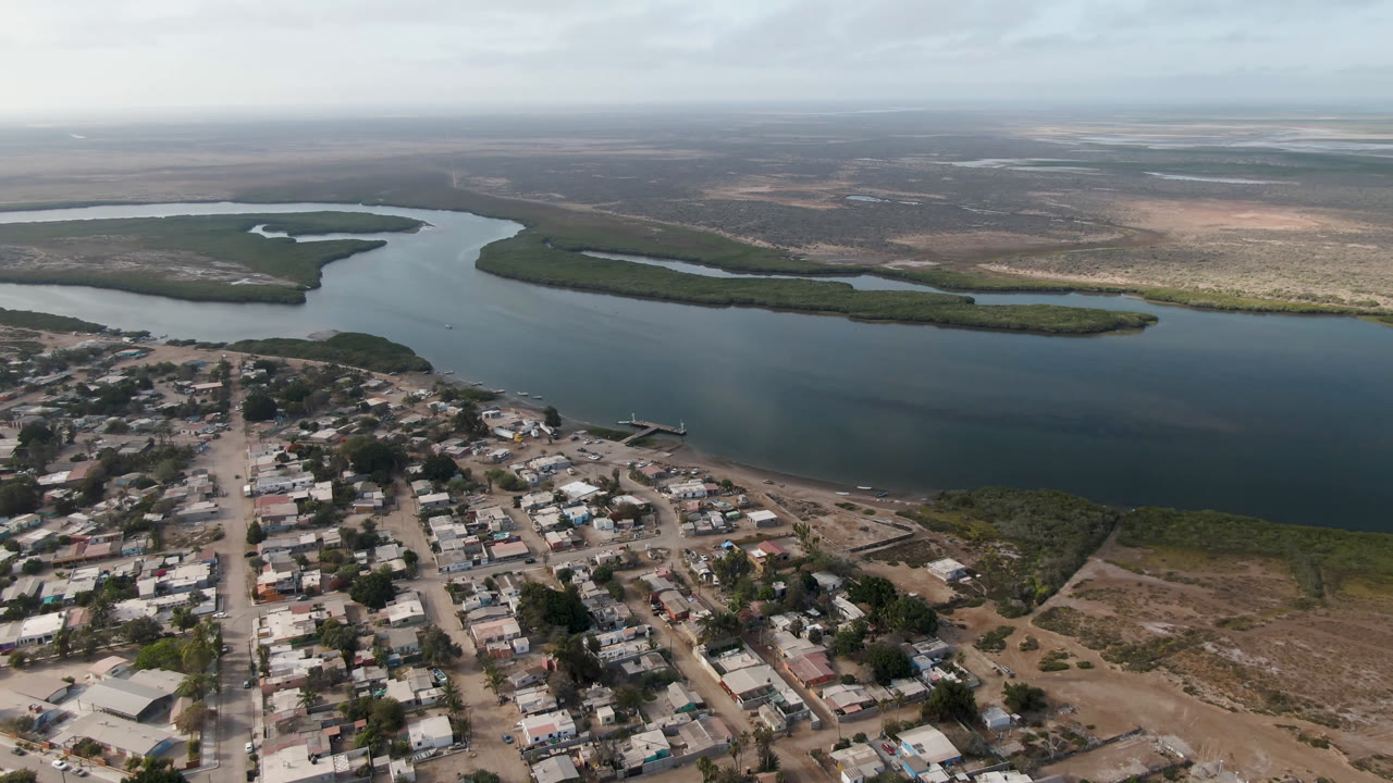Aerial frame from Aldofo Lopez Mateos showing town blocks at the bottom, a small pier reaching toward the water, green riverbank vegetation, distant open land, and no visible people.
