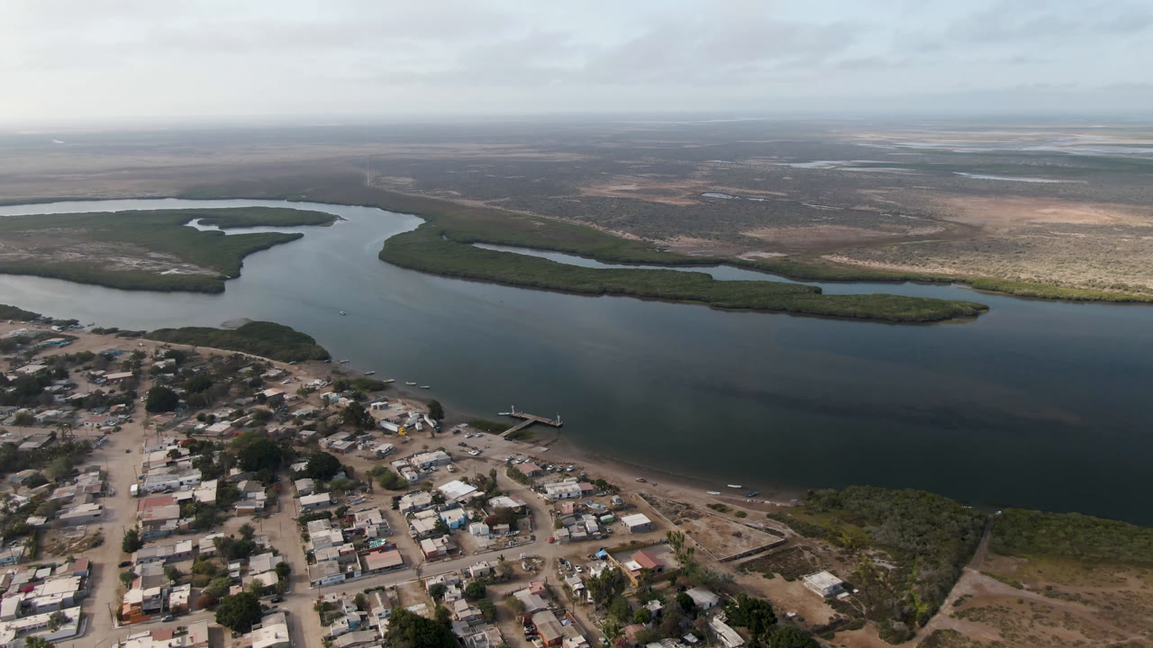 Aerial frame from Aldofo Lopez Mateos showing rooftops and unpaved streets in the foreground, a dark water channel across the center, low green islands, and no visible people.