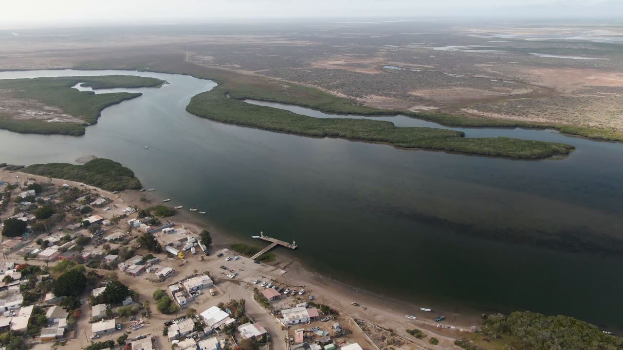 Aerial frame over Aldofo Lopez Mateos, Baja California Sur, with the town edge in the foreground, a dock reaching into the water, tree-covered delta islands, and no visible people.