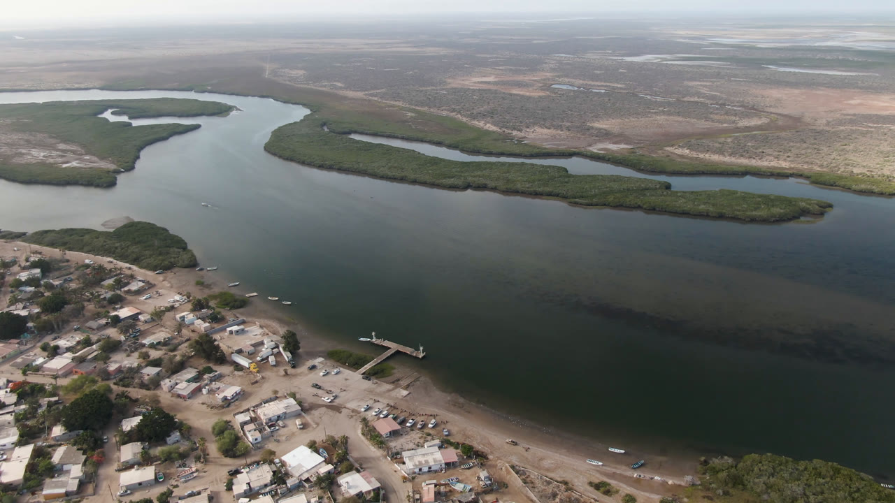 Aerial view in Aldofo Lopez Mateos showing more of the shoreline settlement below, a narrow dock, small boats near shore, green delta vegetation, and no visible people.