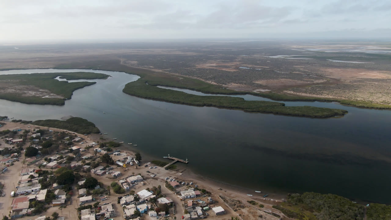 Aerial frame over Aldofo Lopez Mateos showing more of the shoreline settlement in the lower left, a small dock near the water, the wide river or lagoon in the middle, distant scrubland, and no visible people.