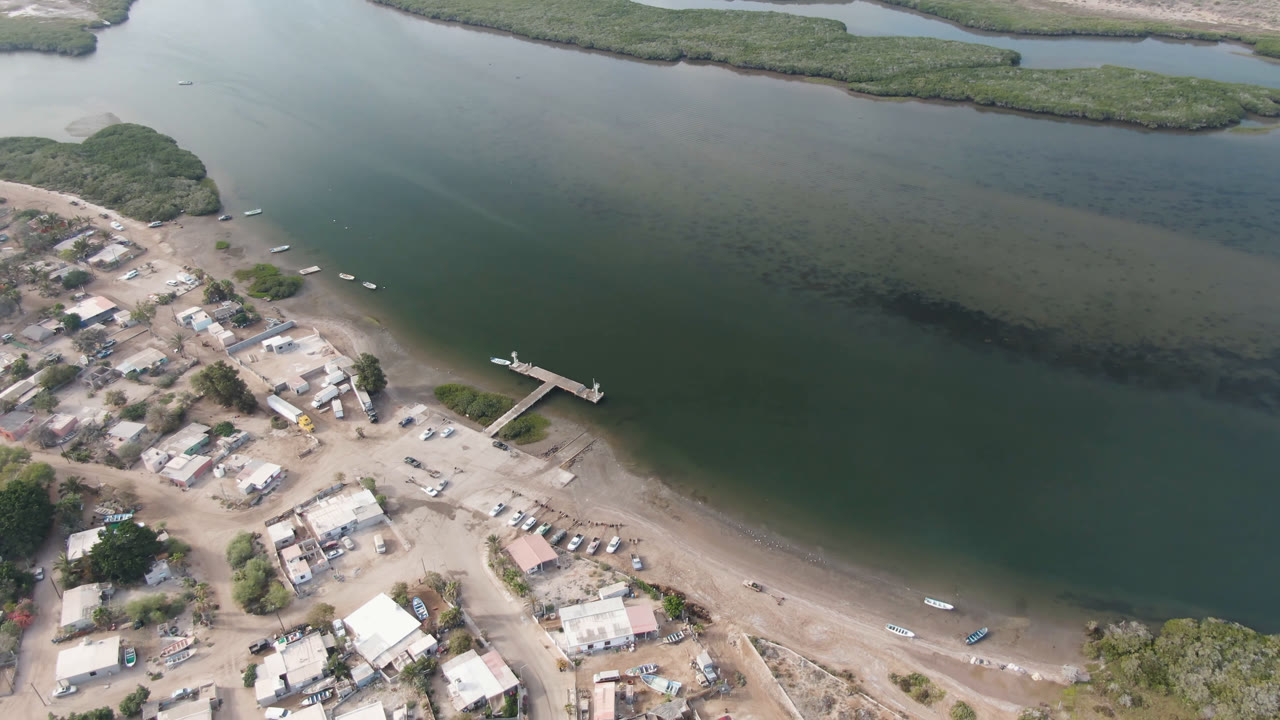 Aerial frame over Aldofo Lopez Mateos, Baja California Sur, showing sandy roads, low buildings, a narrow dock, small boats near shore, calm green water, and no visible people.