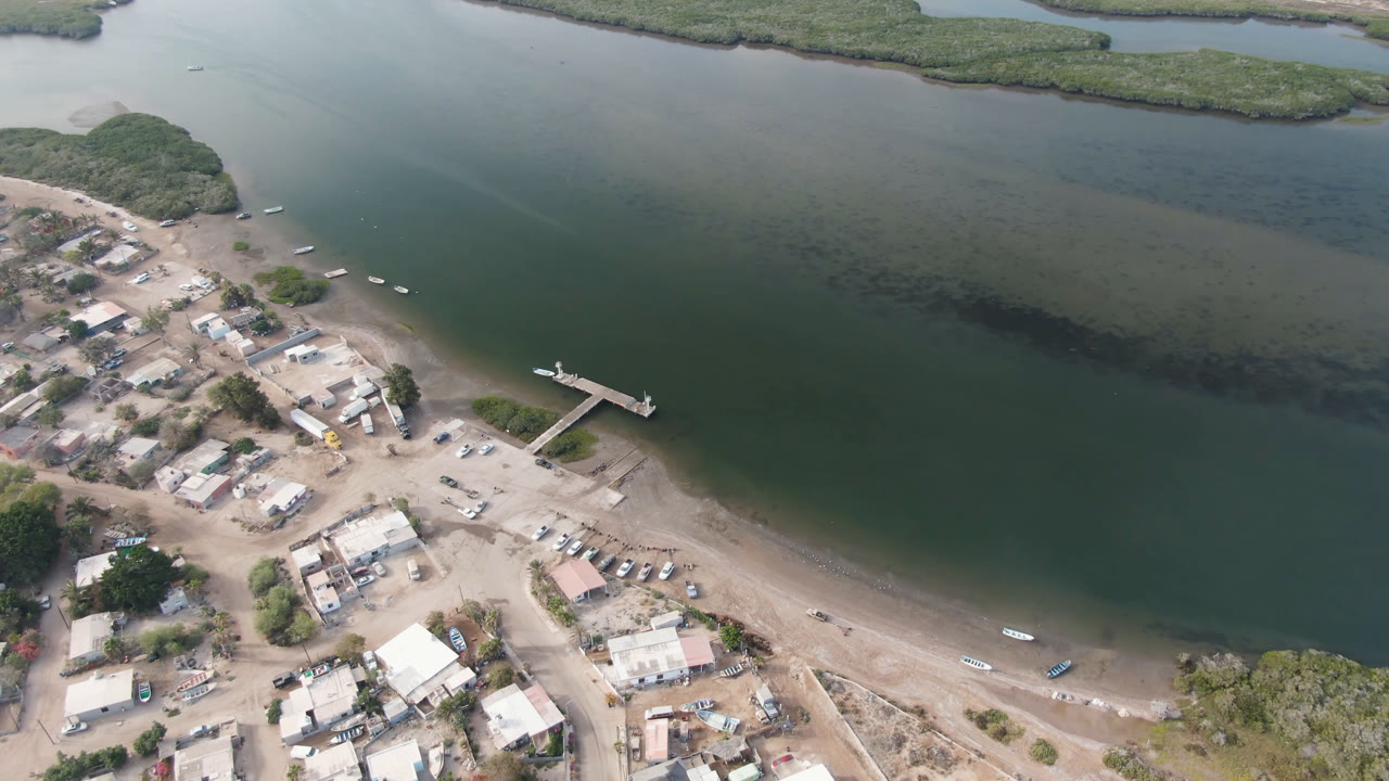 High drone view in Aldofo Lopez Mateos with the town clustered at lower left, a straight dock extending from the beach, small boats along the waterline, and no visible people.