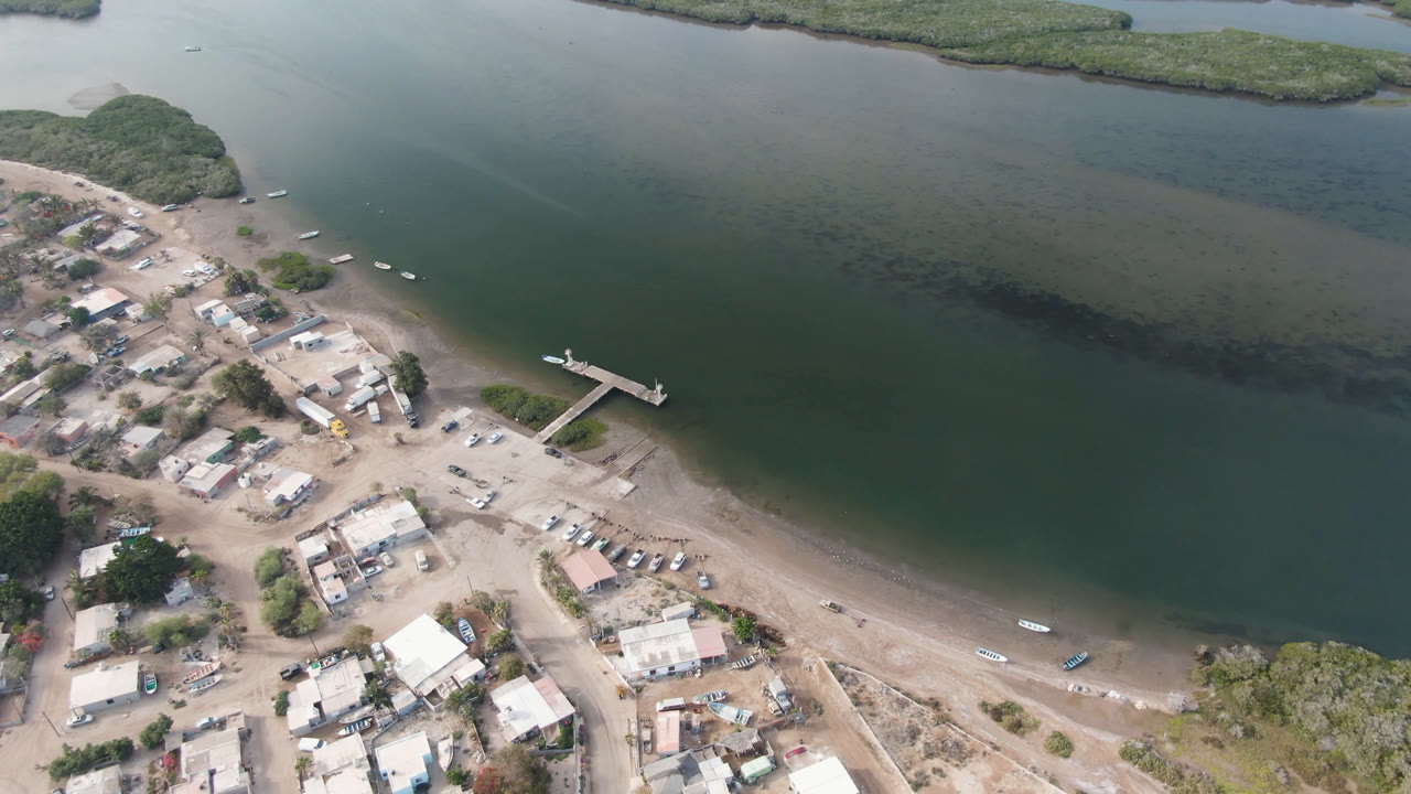 Aerial shoreline scene with a dock, scattered buildings, parked cars, boats, and water channels.