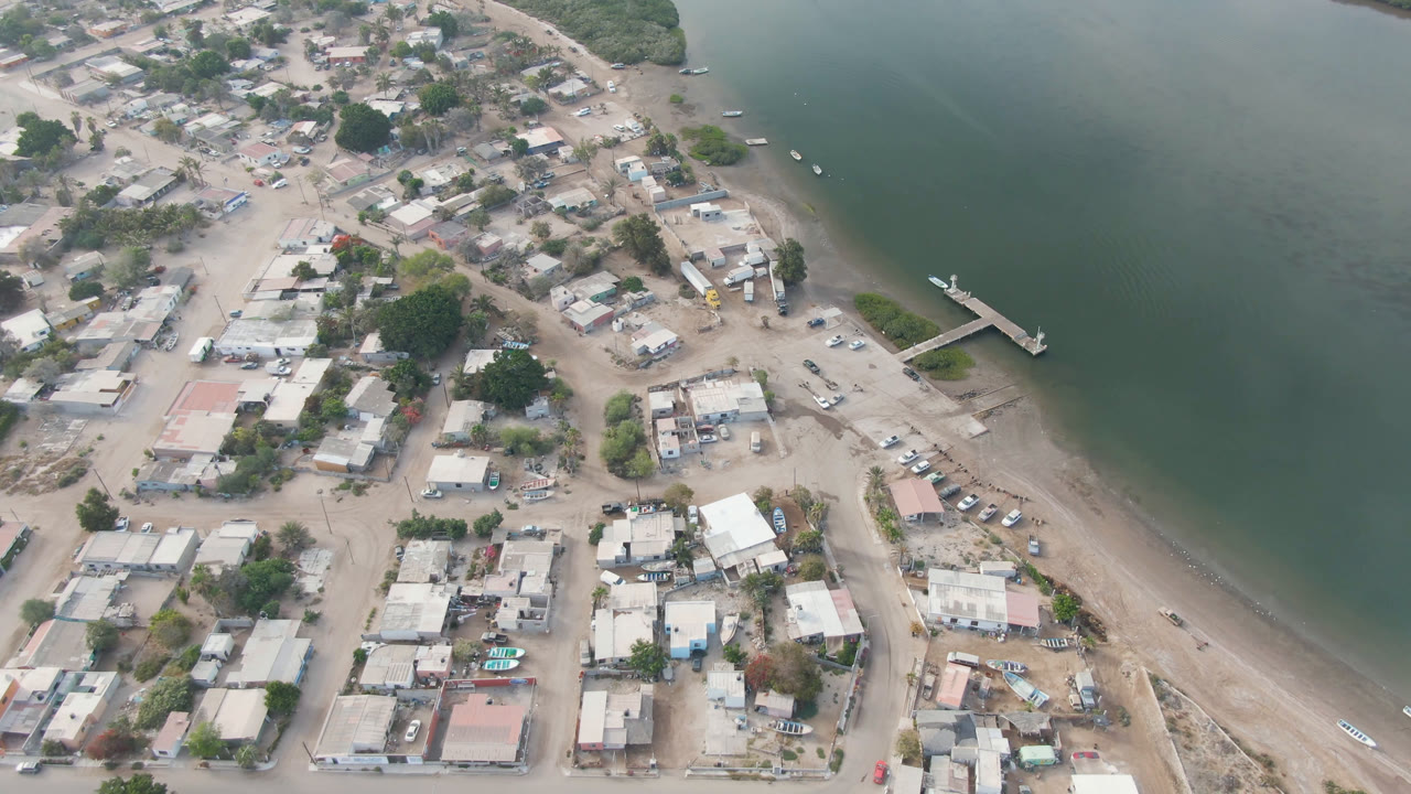 Aerial frame over Adolfo Lopez Mateos with the dock angled into calm green water, town blocks and dirt roads below, vegetation across the channel, and no visible people.