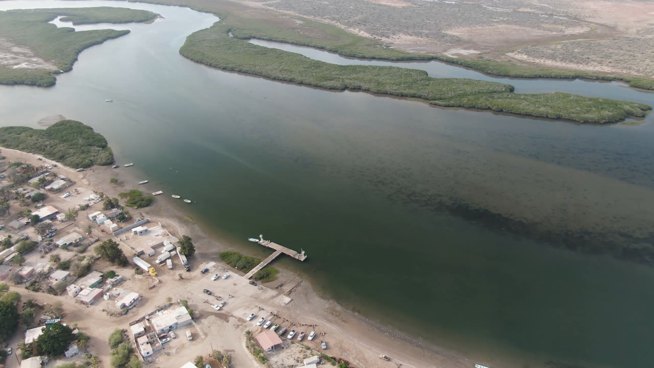 Opening aerial frame over Aldofo Lopez Mateos, Baja California Sur, with green water filling the center, a small dock and buildings along the lower shore, marshy channels in the distance, and no visible people.