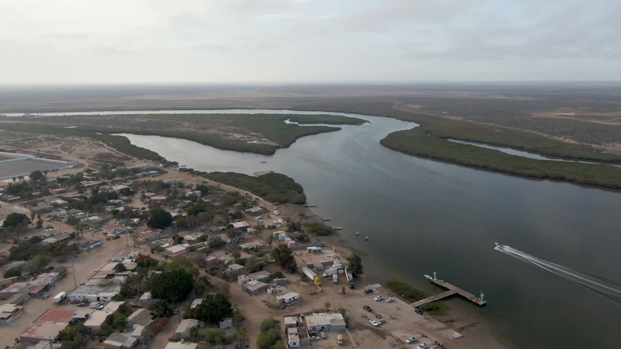 Aerial frame over Aldofo Lopez Mateos showing the waterfront settlement in the foreground, a dock jutting into the river, green marsh banks across the channel, and no visible people.