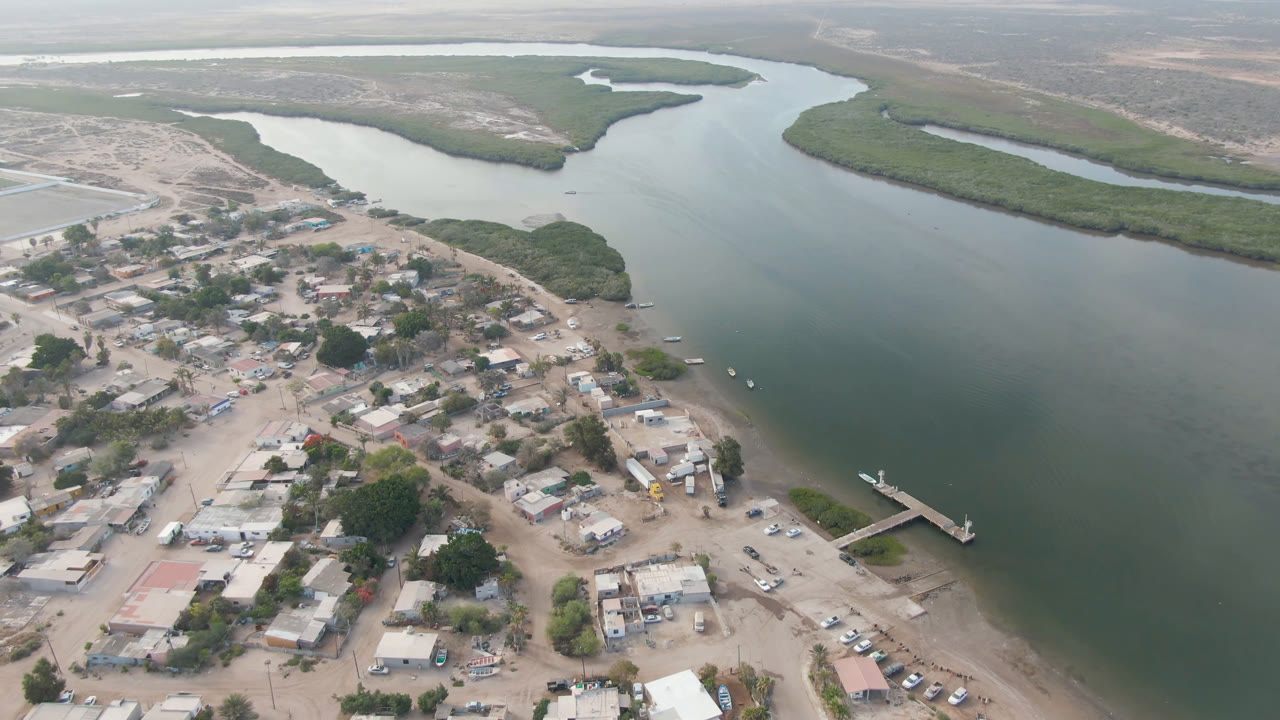 Aerial frame in Aldofo Lopez Mateos showing clustered town buildings in the foreground, a small pier at the river edge, winding water behind it, and no visible people.