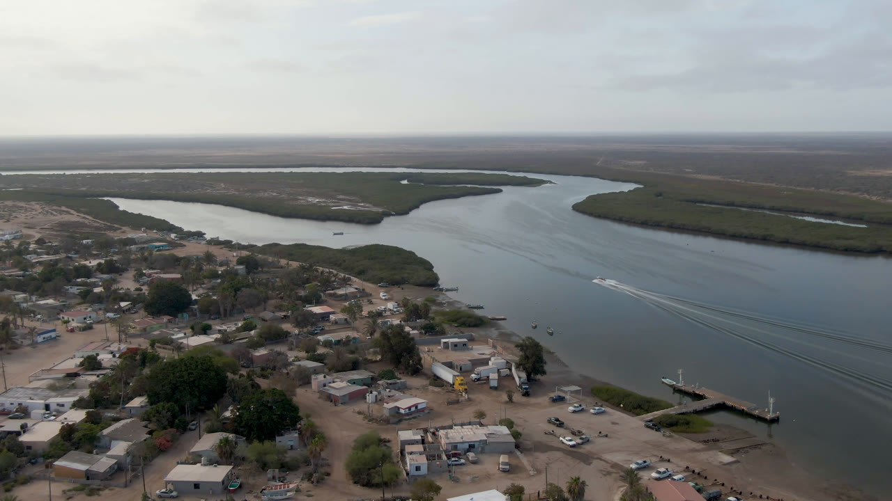 Aerial frame over Aldofo Lopez Mateos with the town in the lower foreground, a pier at the shoreline, the water channel running across the center, and no visible people.
