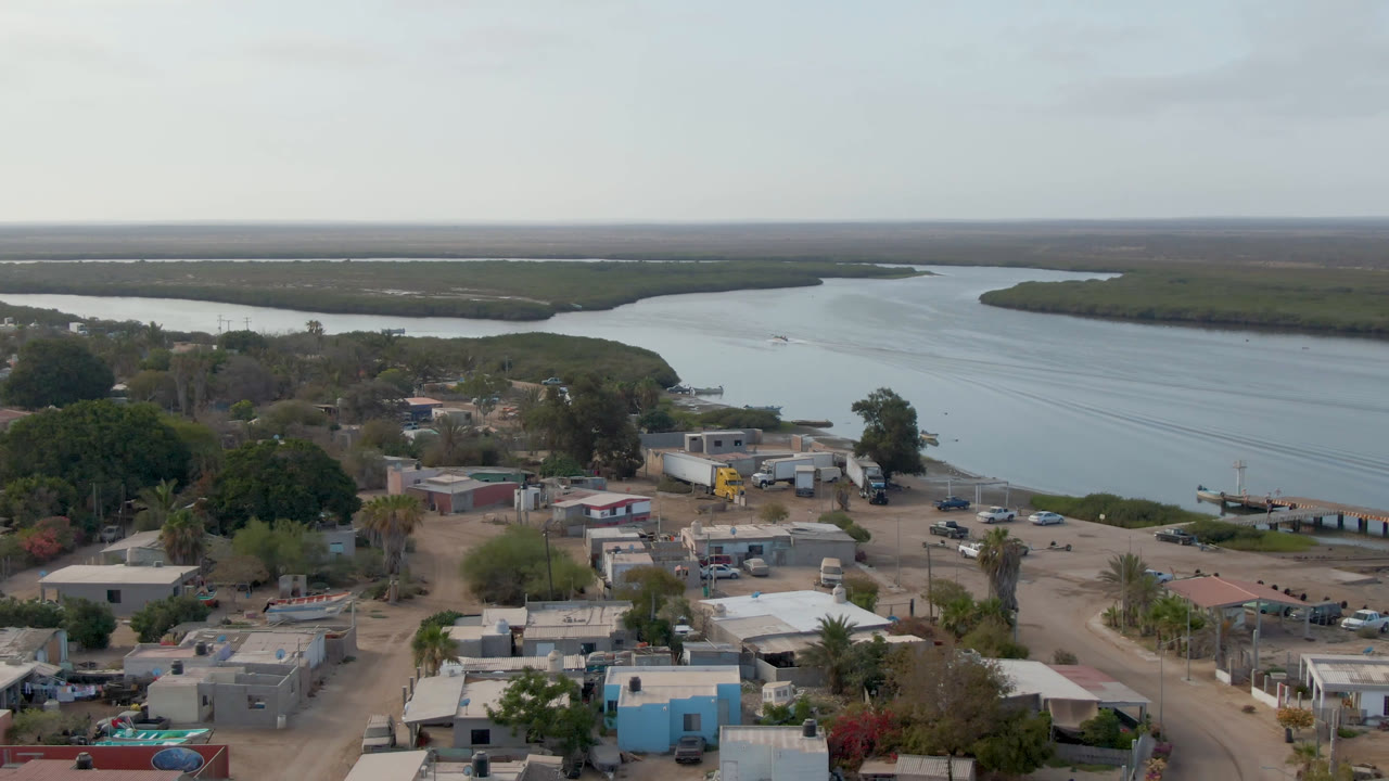 Second aerial frame in Adolfo Lopez Mateos, Mexico, showing compact homes, sandy streets, trees, parked vehicles, waterfront structures, and the river curving behind, with no visible people.