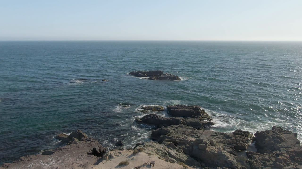 Aerial view over Bahia Ascuncion water with dark rocks offshore, smaller rocks near the lower edge, bright surf lines, clear sky, and no visible people.