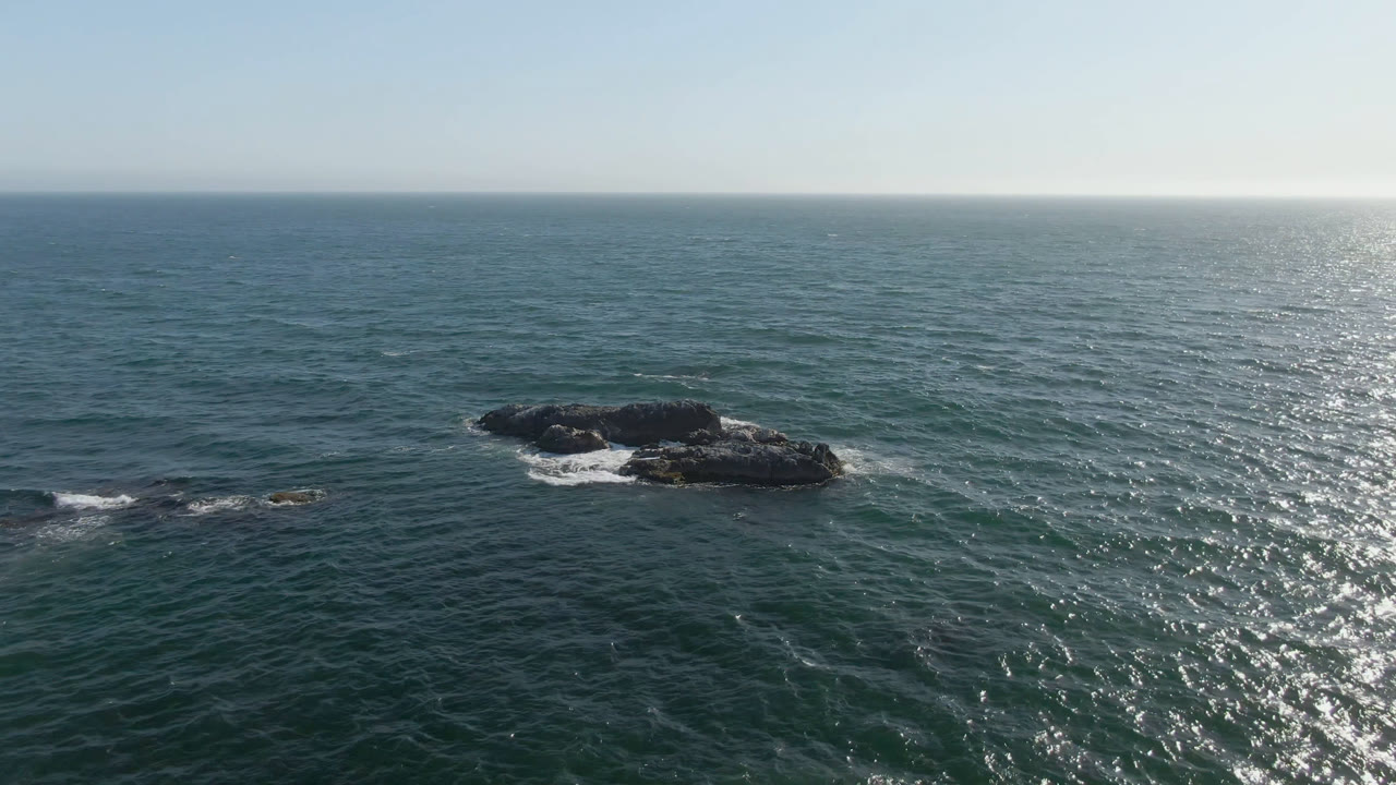 Aerial view in Bahia Ascuncion with offshore rocks near the center, rippled teal water, foam breaking along the rock edges, a pale sky, and no visible people.
