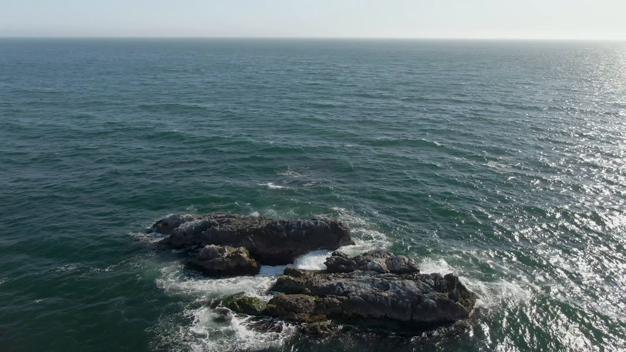Aerial view over Bahia Ascuncion ocean with dark offshore rocks low in frame, white surf around them, sunlit water to the right, and no visible people.