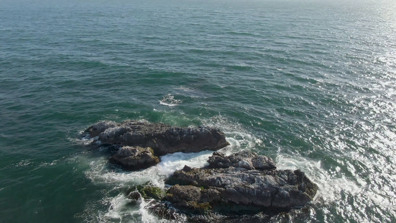 Aerial frame over Bahia Ascuncion showing the main rock formation nearer the center, green-blue water around it, white foam in the channels, and no visible people.
