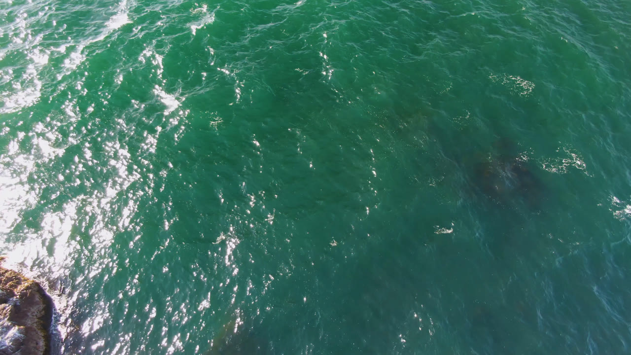 Overhead view of green ocean water in Bahia Ascuncion, Mexico, with bright sun glitter, small foam streaks, and a rocky edge entering the lower left, no visible people.