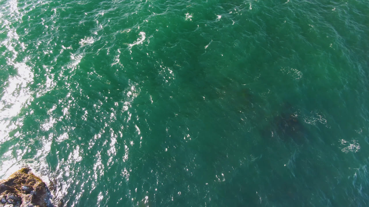 Aerial frame over Bahia Ascuncion green water, where a bright vertical band of reflected sunlight crosses rippled waves and a small rock point appears near the bottom edge, no visible people.