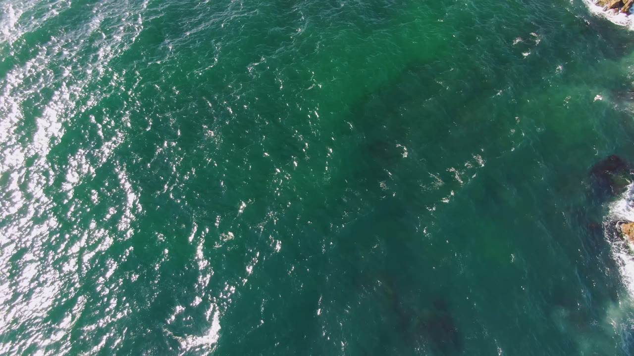 Overhead view of teal water at Bahia Ascuncion with sun glints, white wave texture, and a small rocky edge entering at upper right, with no visible people.