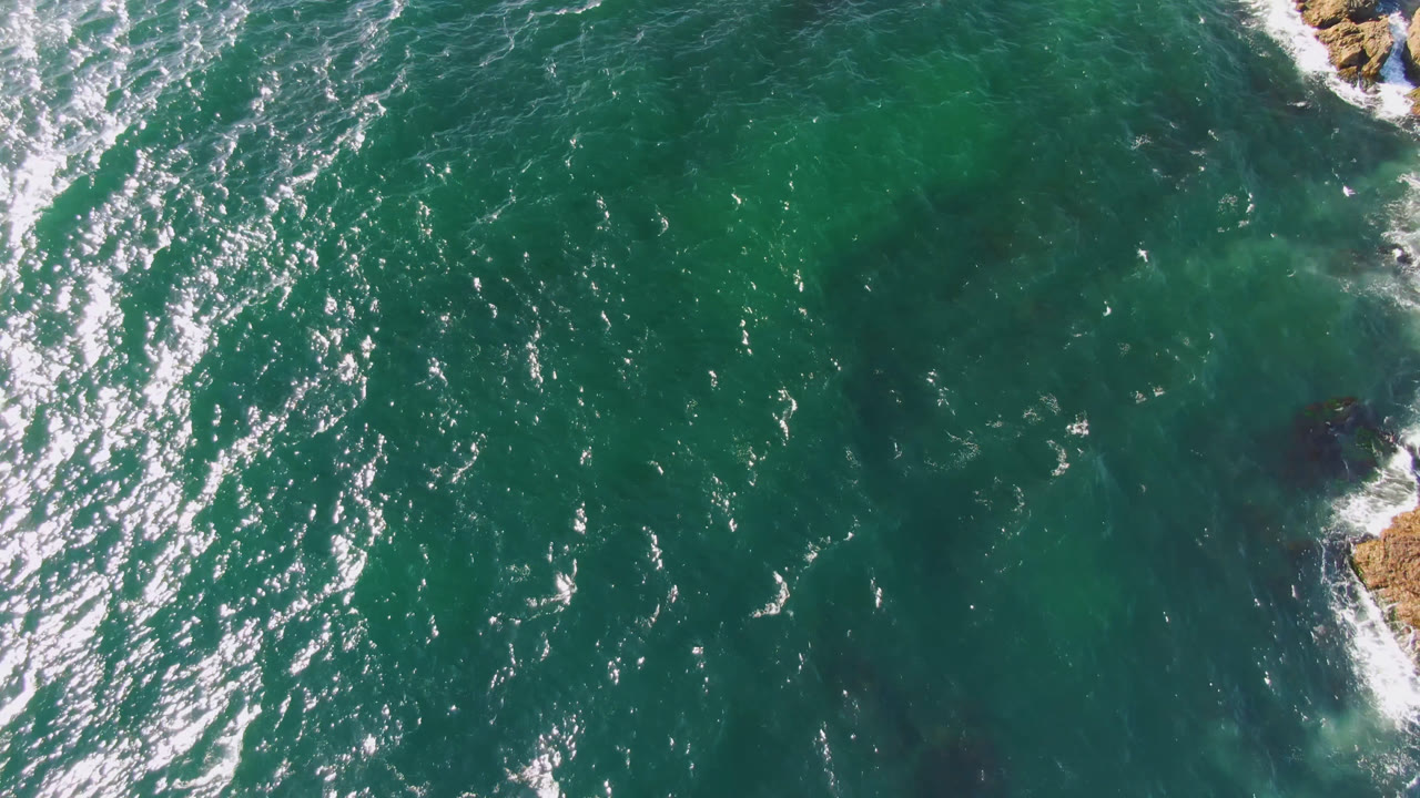 Top-down Bahia Ascuncion ocean frame with bright rippled highlights, dark underwater areas, and a tan rock breaking the water near the right side, with no visible people.