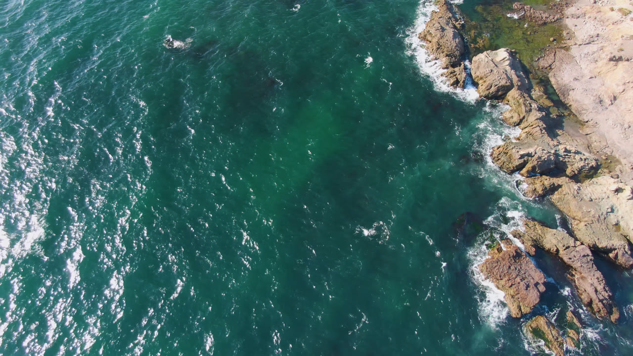 Overhead view of teal water meeting rocky sand at Bahia Ascuncion, Baja California Sur, Mexico, with bright surface reflections and no visible people.
