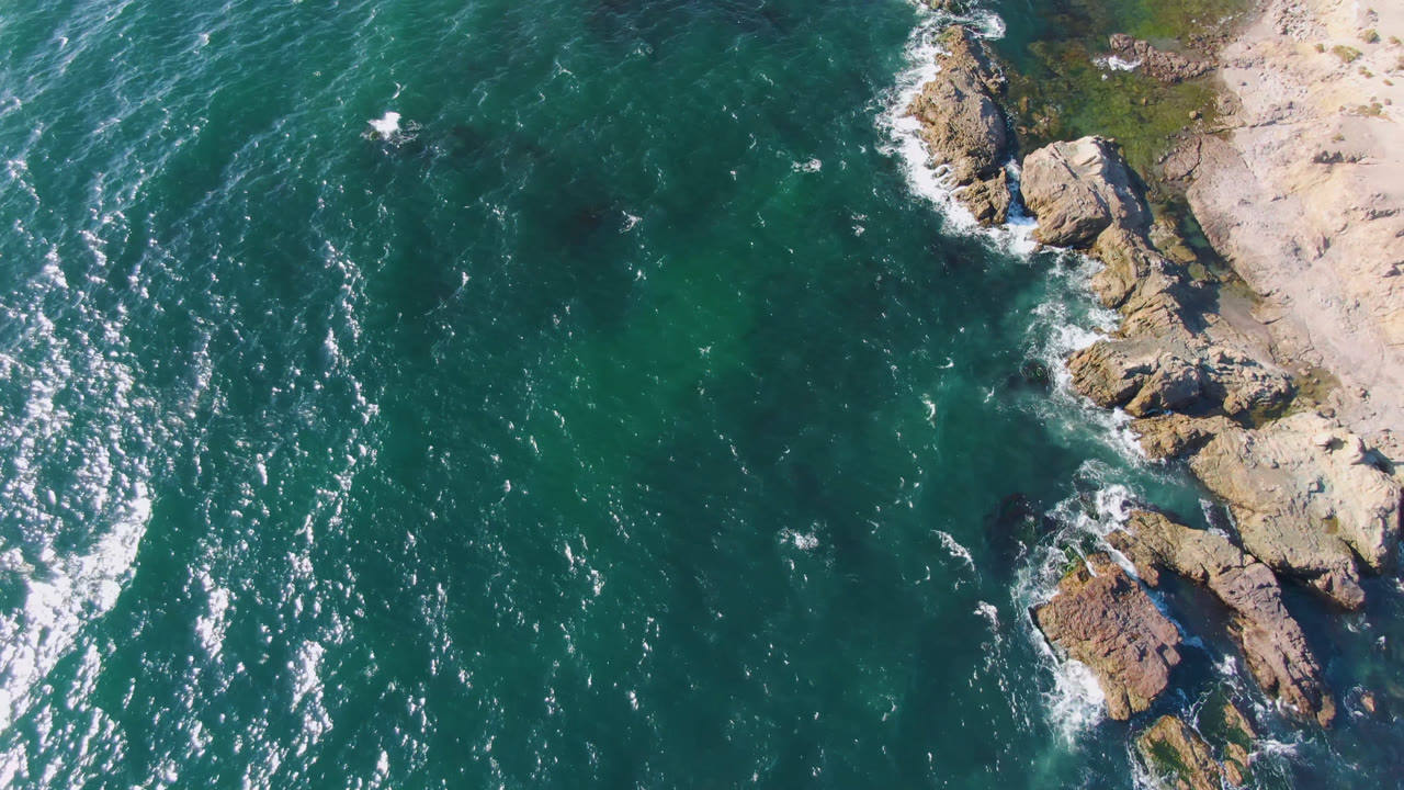 Aerial frame over Bahia Ascuncion showing white foam curling around shoreline rocks, tan beach at the upper right, and no visible people.