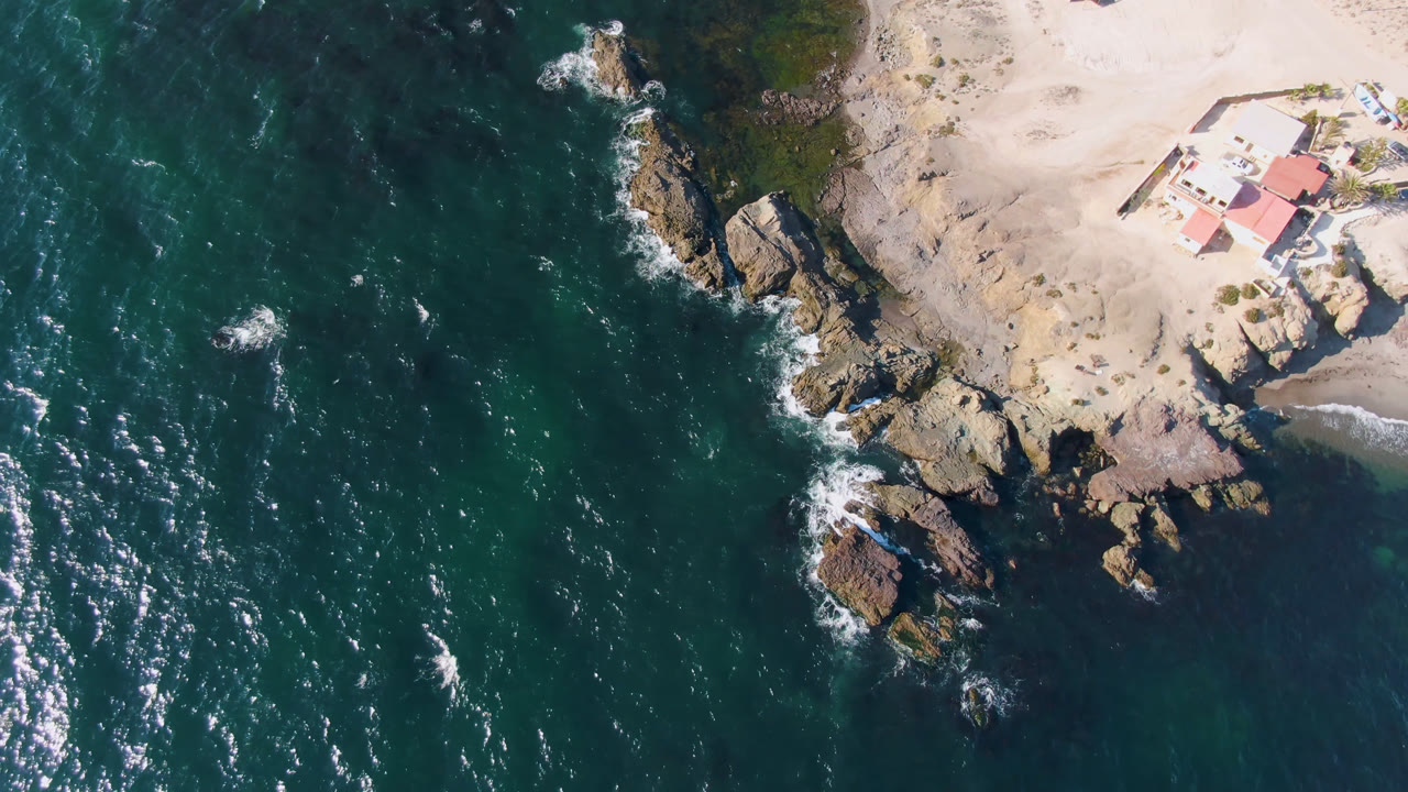 First overhead frame of Bahia Ascuncion coastline with dark teal water on the left, jagged rocks at center, pale sand with a red-roofed house at upper right, a small boat on the beach, and no visible people.