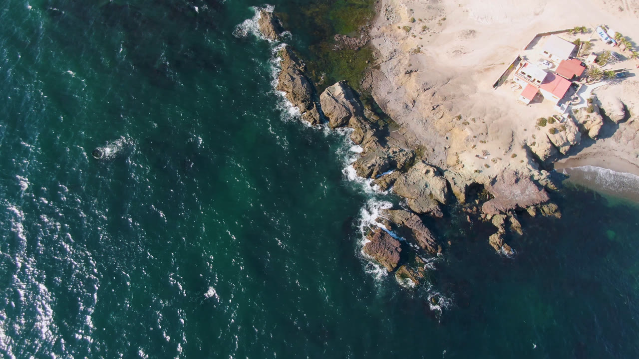 At 0:01, the Bahia Ascuncion aerial shows more white wave streaks in the water, with the rocky point below the sandy beach, the red-roofed house near the top right, the small boat on shore, and no visible people.