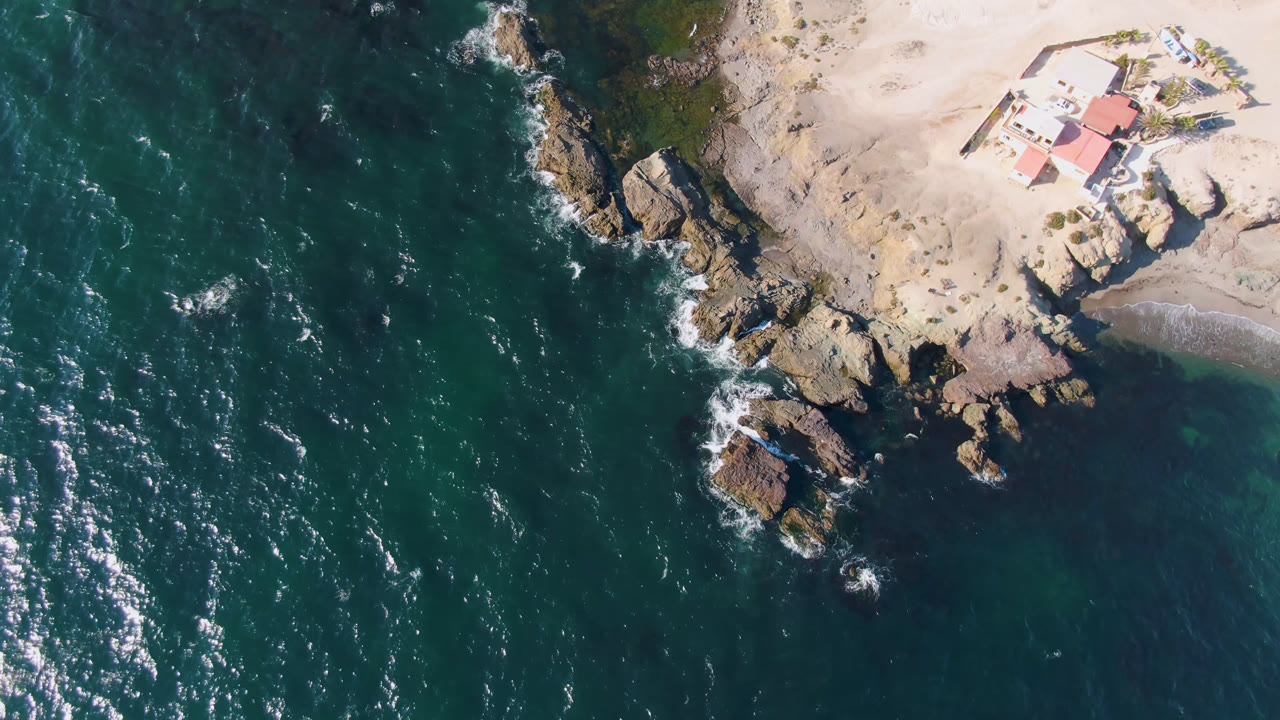 Top-down coastline with teal water, surf, rocks, sandy beach, house, and boat.