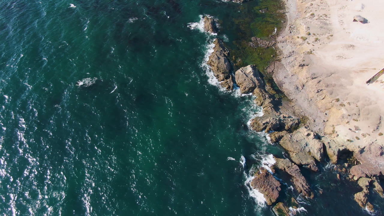 Aerial view of dark turquoise ocean beside pale sand and coastal rocks.