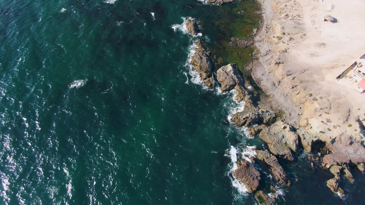 Overhead view near Bahia Ascuncion shows ocean filling the left side, a sandy beach edge on the right, scattered rocks, and no visible people.