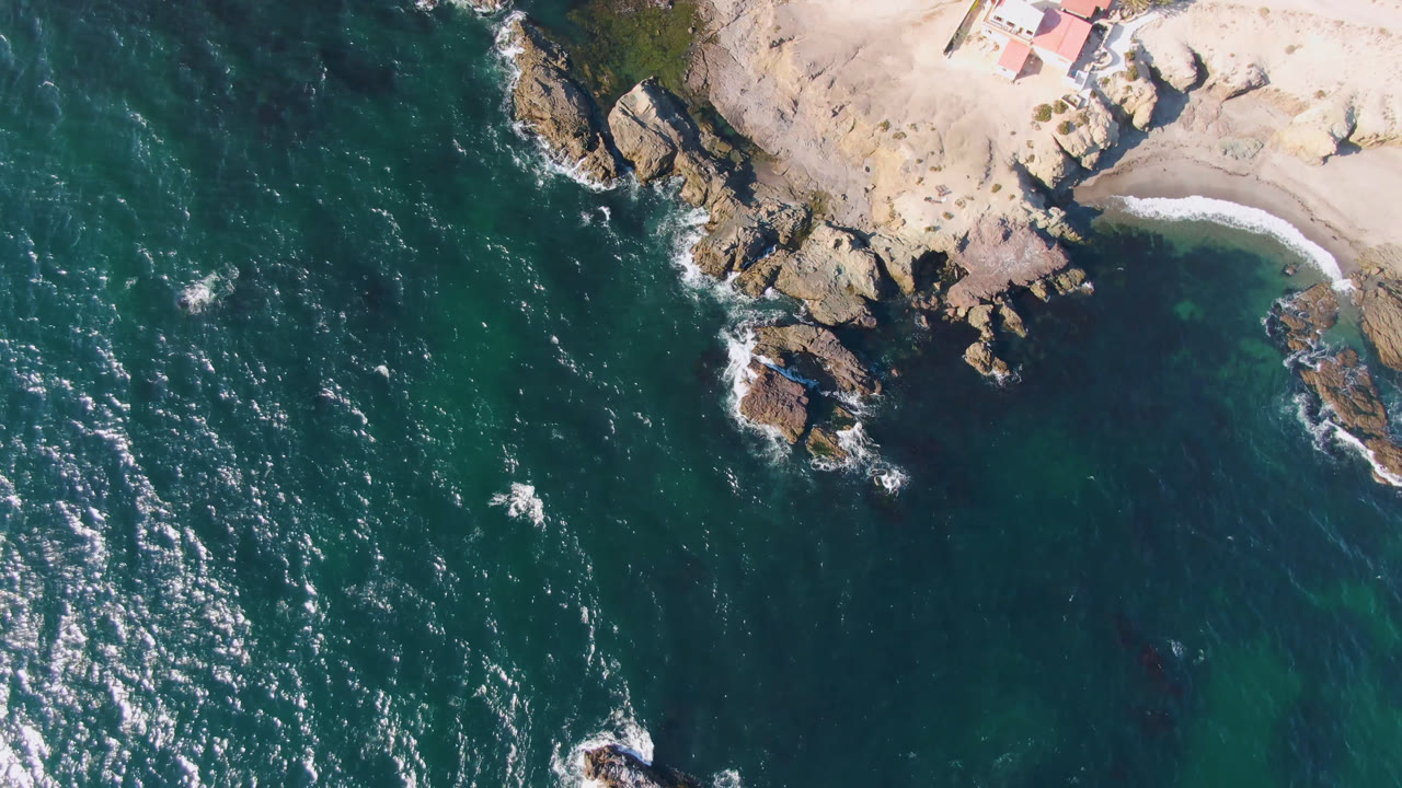 Overhead drone frame of Bahia Ascuncion showing teal ocean, pale rocks, a small sandy cove, and no visible people at the start of the clip.
