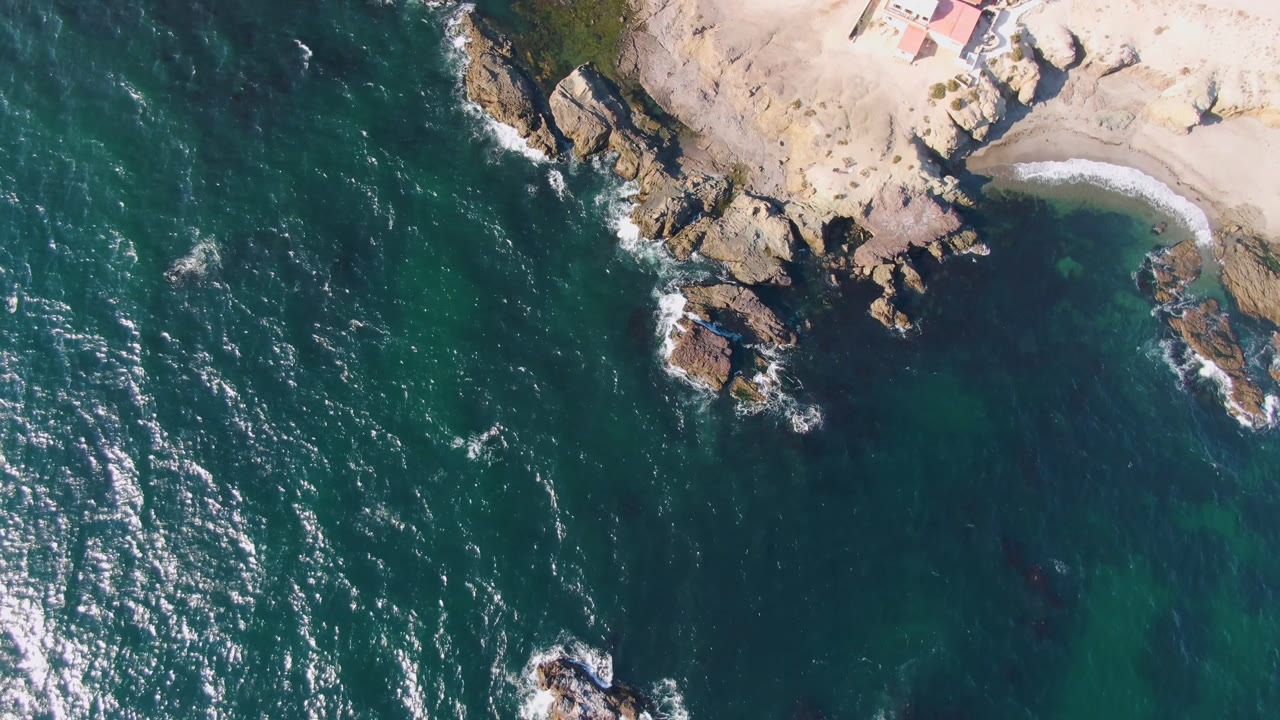 Aerial view over Bahia Ascuncion with bright sun reflections on dark teal water, tan shoreline rocks, a curved beach, and no visible people.