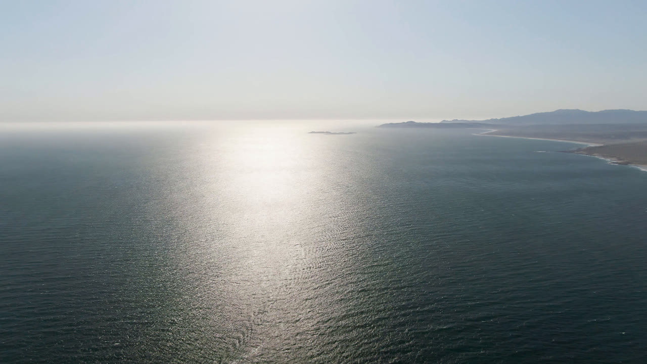 Wide aerial frame over Bahia Ascuncion water in Baja California Sur, with bright sun reflection on the ocean, a distant coastline at right, low mountains on the horizon, and no visible people.