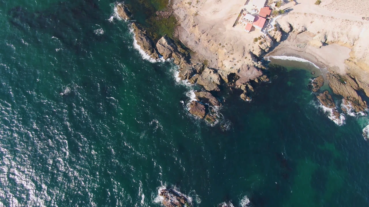 Overhead frame from Bahia Ascuncion, Baja California Sur showing dark teal ocean, wave foam around rocks, pale sand, and a red-roofed coastal house at upper right with no visible people.