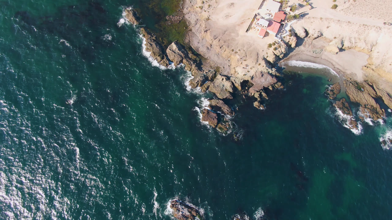 Drone view over Bahia Ascuncion with bright sun flecks on the water, surf curling around central rocks, sandy shoreline above, and the house still visible at upper right with no visible people.
